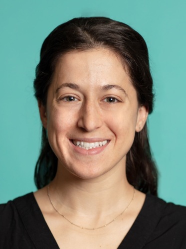 A woman with long dark hair wearing a black shirt and a thin necklace, smiles at the camera against a teal background.